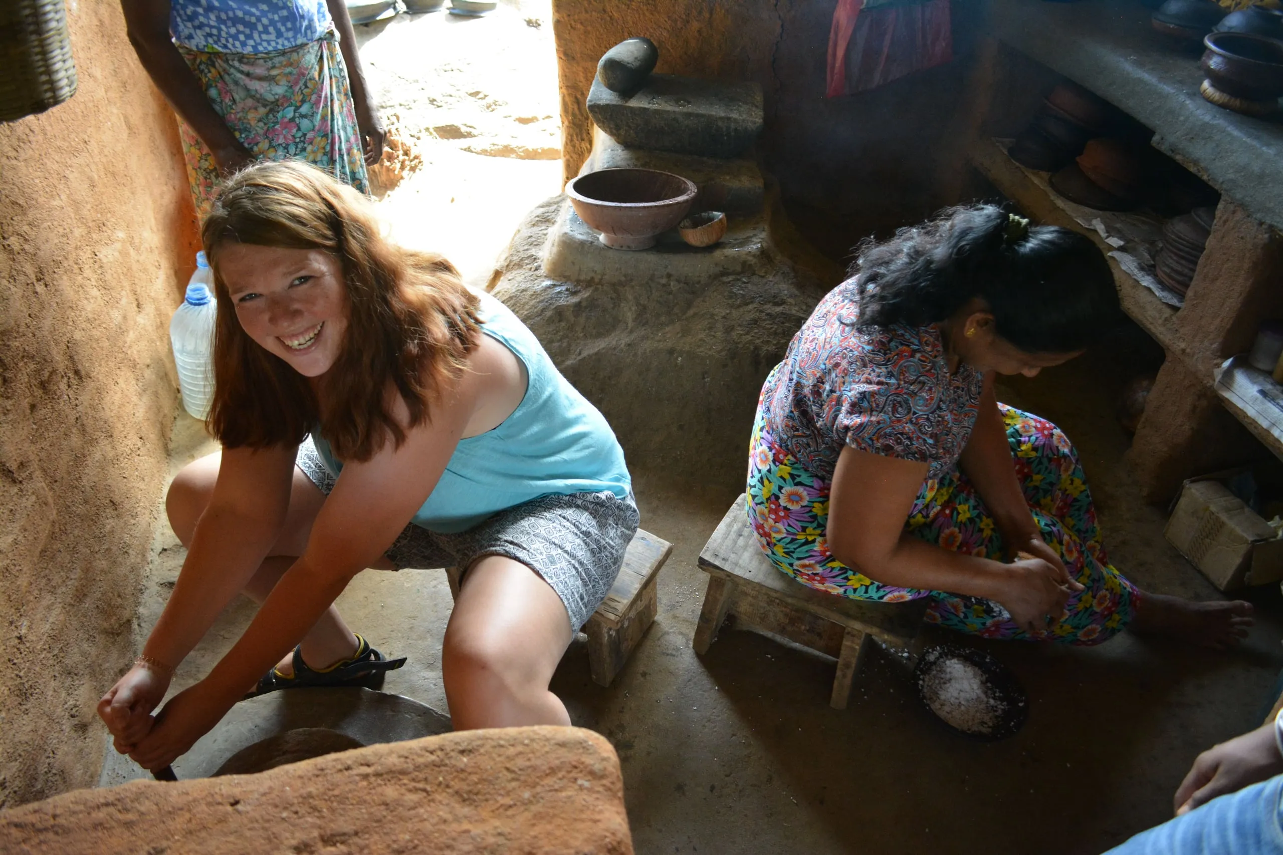 A guest grating coconut on a low stool beside a village woman in a clay kitchen